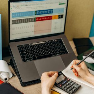 Hands writing notes beside a laptop displaying data in a bright office setup.