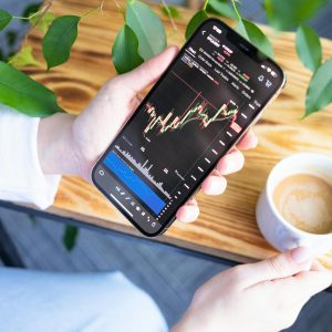 Woman holding smartphone showing stock market graph while enjoying a cup of coffee.