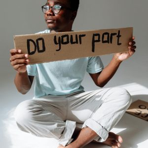A young man holding a cardboard sign reading 'Do Your Part' while sitting indoors.